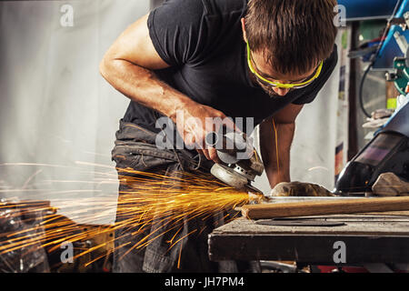 A young man is a brunette welder wearing a black T-shirt and safety goggles grinding metal with a angle grinder in the workshop, sparks flying in the  Stock Photo