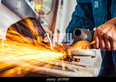 Close-up on the sides fly bright sparks from the welding machine. A young male welder in a blue working overall and working gloves grinds a metal prod Stock Photo