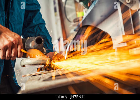 Close-up on the sides fly bright sparks from the welding machine. A young male welder in a blue working overall and working gloves grinds a metal prod Stock Photo