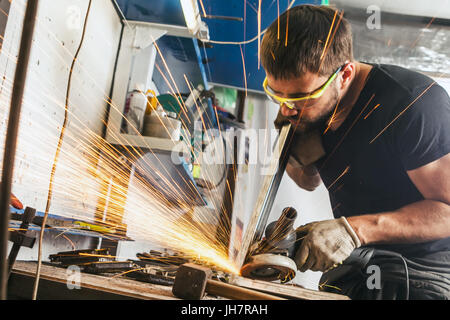 A young brunette man welder in a black T-shirt and safety glasses polishes the metal angle grinder in the workshop, sideways flashes Stock Photo
