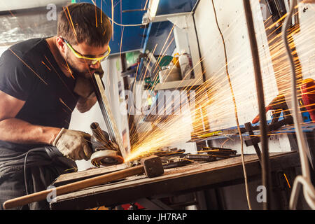 A young brunette man welder in a black T-shirt and safety glasses polishes the metal angle grinder in the workshop, sideways flashes Stock Photo