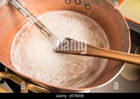 Factory of the production of delicious caramel candies. First step. Boiling syrup of sugar in copper pot with thermometer and big spoon. White colour  Stock Photo