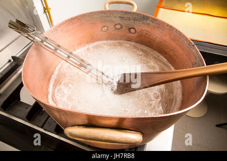 Factory of the production of delicious caramel candies. First step. Boiling syrup of sugar in copper pot with thermometer and big spoon. White colour  Stock Photo