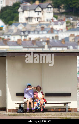 A couple sitting togother under the shade of a promenade shelter at the coastal resort town of Llandudno in North Wales with the Great Orme and town in the distance Stock Photo