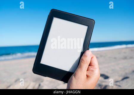 closeup of the hand of a young caucasian man on the beach holding a tablet or e-reader with a white blank space in its screen Stock Photo