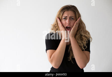 Young, blonde, beautiful, milennial woman expressing excitement and confusion against a white background and in a black shirt indoors during the day. Stock Photo