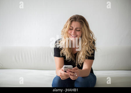 Young, blonde, beautiful milennial woman expressing happiness while playing on her smartphone, sitting on a couch against a white background. Stock Photo