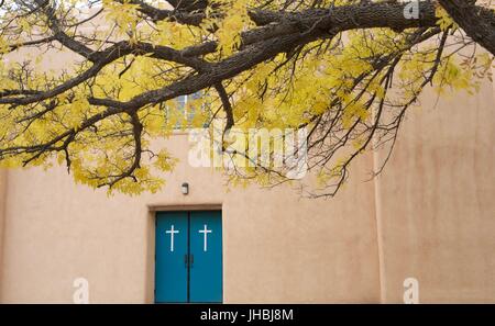 Beautiful tree with yellow leaves outside of adobe church with turquoise doors with white crosses on them Stock Photo