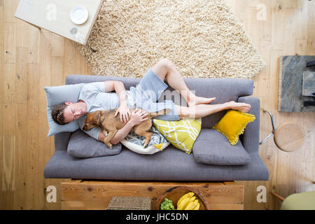 Man is lying on the sofa in the living room of his home. He has his dog lying on him who is also asleep. Stock Photo