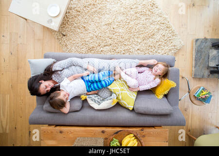 A mother is lying on the sofa in the living room of her home, with her son and daughter. The girl is lying on the other end of the sofa and is laughin Stock Photo