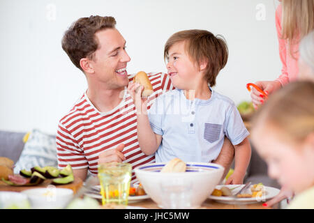 Little boy is sitting on his dads lap at the dining table in their home at dinner time. He is trying to feed his father a bread bun. Stock Photo