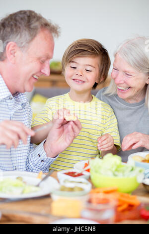 Little boy is sitting at the dining table with his grandparents in their home. His grandfather is buttering some bread and giving it to his grandson. Stock Photo