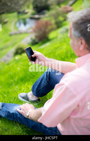 Mature man is sitting on the grass in his garden while using a smartphone. Stock Photo