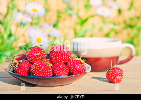 warm cross-processed image of red ripe strawberries in plate on wooden table in garden with tea cup and camomile flowers at background on sunny summer Stock Photo