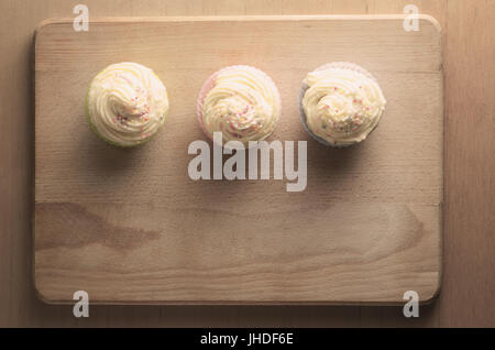 Overhead shot of three cupcakes in a row on a wooden chopping board, topped with swirls of buttercream icing and colourful sprinkles. Retro hues. Stock Photo