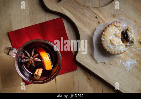 Overhead shot of a glass of mulled wine on red napkin, with partially eaten mince pie on wooden paddle board.  Light wood table below. Stock Photo