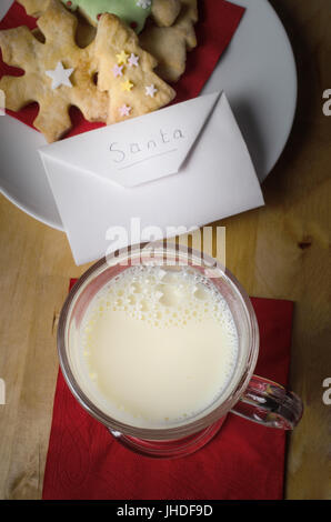 Christmas night scene with a plate of  decorated biscuits (cookies) and glass of milk with envelope addressed to Santa on wooden table. Stock Photo