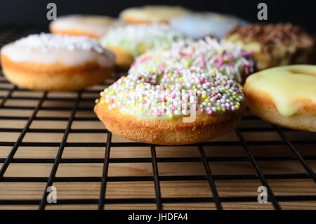 Home baked mini doughnut sponge cakes, with a variety of decorative sprinkles, on a black wire cooling rack and wooden table. Stock Photo