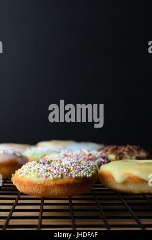 Mini doughnut cakes, decorated with a variety of sprinkles, on a black wire cooling rack with wooden table beneath.  Black chalkboard background provi