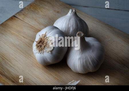 Still life grouping of three whole garlic bulbs on an old, light wood chopping board with painted white planked kitchen table visible below. Stock Photo