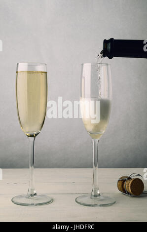 Sparkling white wine is pouring from a bottle into one of two champagne flutes on an old painted wood plank table. The second glass is already full. W Stock Photo