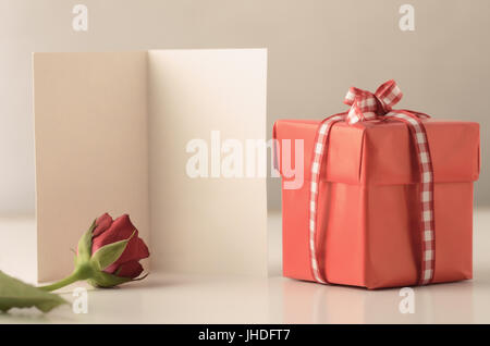 A single small square gift box with lid, tied to a bow with gingham ribbon on a white table with a rose and an opened card providing copy space. Stock Photo