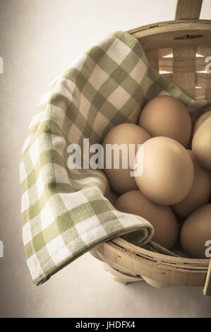 A basket of gathered eggs lined with green and white checked gingham cotton cloth.  Partly desaturated and vignetted for retro, vintage effect. Angled Stock Photo