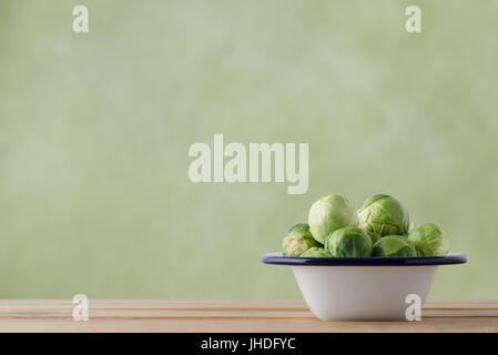 Enamel cooking tin filled with fresh, raw brussel sprouts on light wood plank table.  Green background provides copy space. Stock Photo