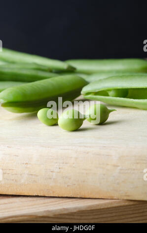 Close up of three individual peas on wooden chopping board, freshly picked from group of pods behind. Stock Photo