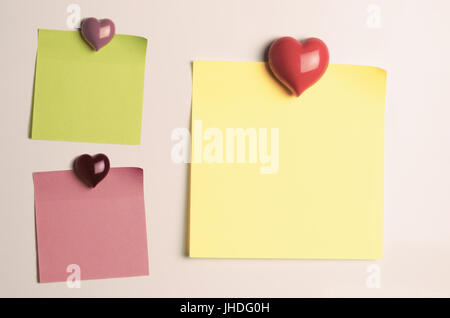 Three sticky note paper squares in green, pink and yellow, stuck to fridge door with heart shaped magnets. Left blank to provide copy space. Stock Photo