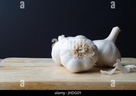 Three whole garlic bulbs on old light wood chopping board with scattered peelings. Black chalkboard background. Stock Photo