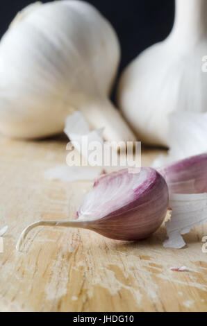 Close up of a garlic clove on an old wooden chopping board with two whole bulbs and peelings in soft focus behind. Black background. Stock Photo