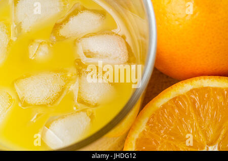 Angled overhead close up shot of fresh juice in glass with ice cubes, beside whole and half oranges on wooden table. Stock Photo