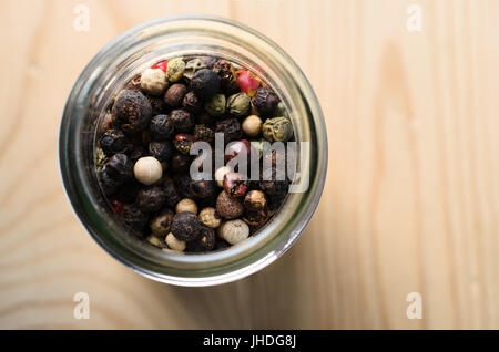 Overhead close up shot of mixed peppercorns in a glass container on a light wood table with copy space to the right. Stock Photo