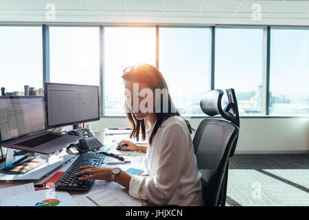 Woman entrepreneur working in her office with computers and business papers on table. Young woman operating computer while looking at business papers. Stock Photo