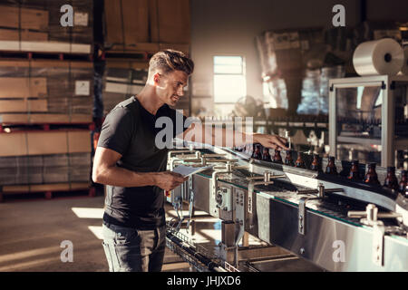 Young man supervising the beer production at brewery. Man working at alcohol manufacturing factory. His is standing near a bottling machine. Stock Photo