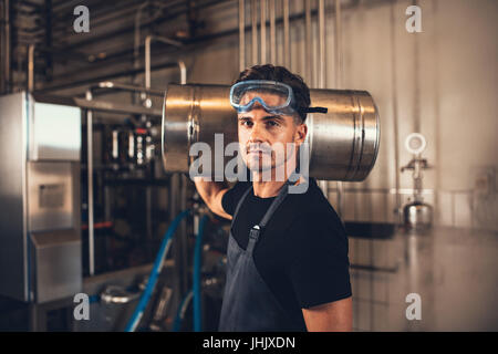 Portrait of male brewer with keg at brewery factory. Young man holding a metal beer container on shoulder at warehouse. Stock Photo