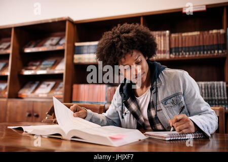 University student taking notes from book while sitting in the library. Young black man writing notes in library. Stock Photo