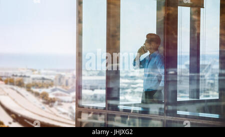 Businessman standing by window and talking on mobile phone. Man standing inside office building and using cell phone. Stock Photo