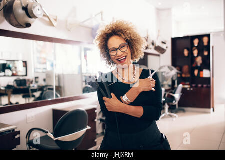 Smiling female hairdresser holding a hair straightener and scissors. Well equipped beauty salon with professional hairdresser. Stock Photo