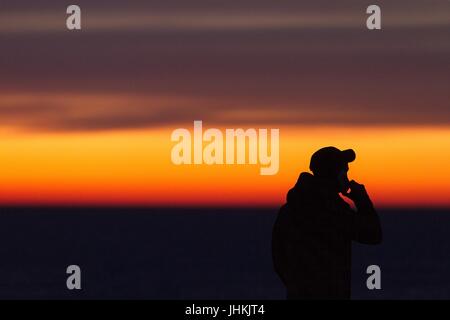 Silhouette of the man with mobile/cellphone during sunset at the seaside. Man in a baseball cap. Stock Photo