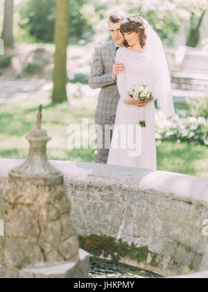 The groom is hugging the bride back while standing behind the fountain. Stock Photo