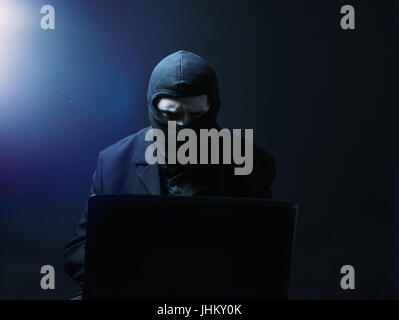 Angry computer hacker in suit stealing data from laptop in front of black background and blue light Stock Photo