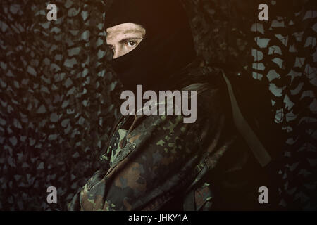 army soldier in uniform with dirty face and balaclava looking sideways in front of camouflage net Stock Photo