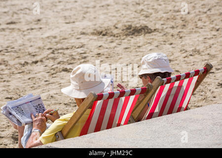 Couple sitting in deckchairs on the beach doing crossword and looking at magazine at Lyme Regis, Dorset in July Stock Photo