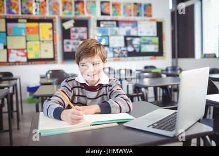 Portrait of smiling elementary schoolboy studying while sitting at desk in classroom Stock Photo