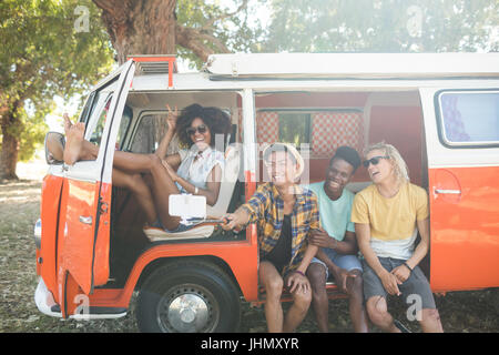 Young friends taking selfie through monopod while sitting together at campsite Stock Photo