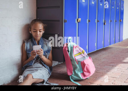Elementary student listening music through headphones while using mobile phone by lockers at school Stock Photo