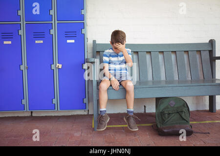 Full length of sad boy sitting on bench by lockers at school Stock Photo