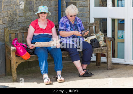 Two mature women sitting on bench, one holding the Lyme Regis Guide, the other getting suntan lotion out of bottle, at Lyme Regis, Dorset in July Stock Photo